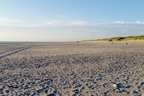 Ein weiter Sandstrand mit Fußabdrücken, die zum Wasser führen, einer Düne im Hintergrund und einem klaren Himmel.