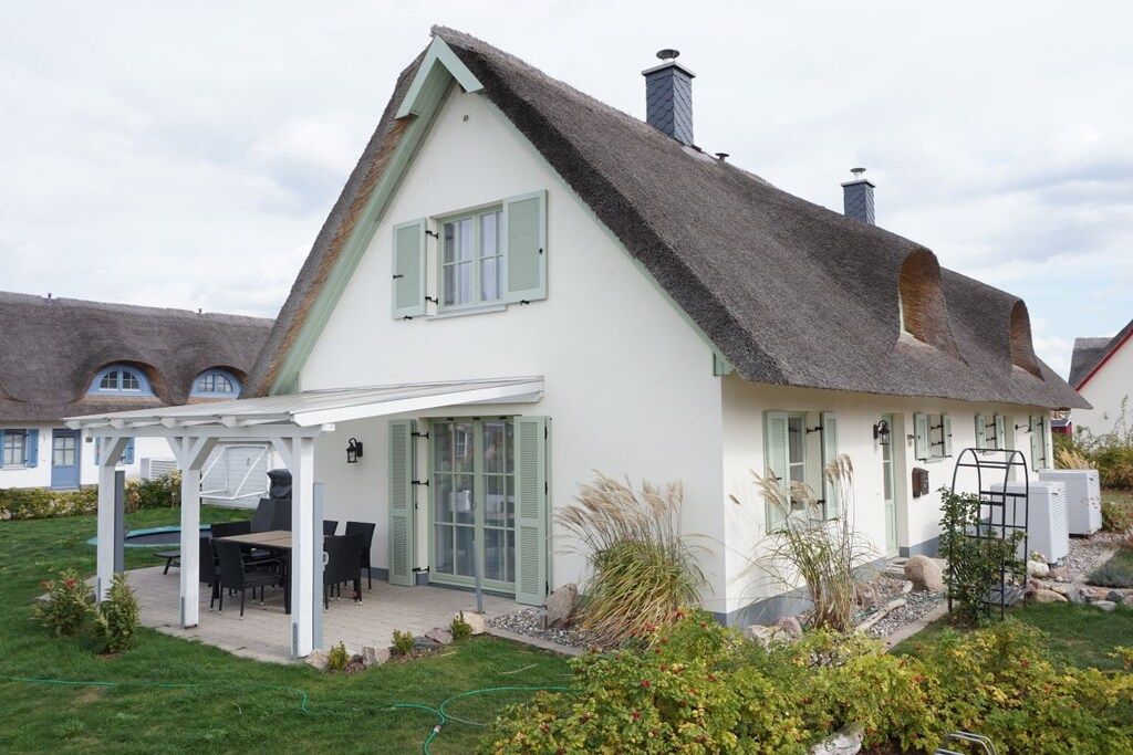 Ein Haus mit weißer Fassade, grünen Fensterläden, einem Strohdach und einer überdachten Terrasse mit Gartenmöbeln.