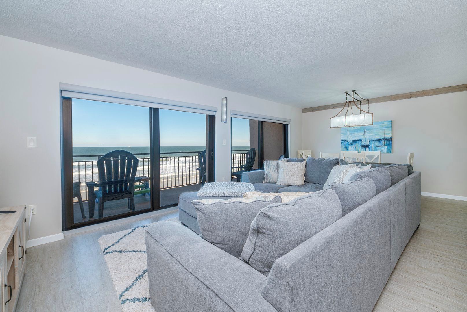 A modern living room with a grey sectional sofa, a large sliding glass door leading to a balcony, and a painting on the wall.