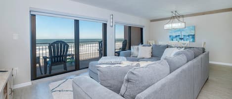A modern living room with a grey sectional sofa, a large sliding glass door leading to a balcony, and a painting on the wall.