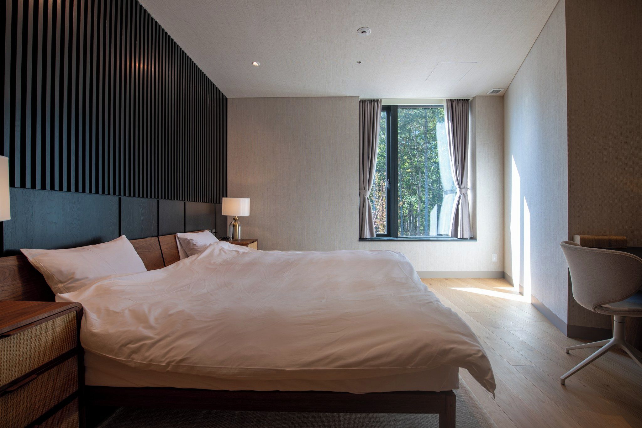 Quarto de hotel moderno com uma cama grande, uma mesinha de cabeceira com abajur, uma cadeira e uma janela com cortinas.