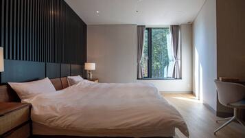 Quarto de hotel moderno com uma cama grande, uma mesinha de cabeceira com abajur, uma cadeira e uma janela com cortinas.