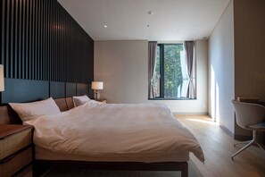 Quarto de hotel moderno com uma cama grande, uma mesinha de cabeceira com abajur, uma cadeira e uma janela com cortinas.