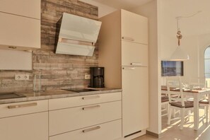 A modern kitchen with a built-in stove, a coffee maker, and a dining area with white chairs and a table. - Apartment Große Freiheit (Ostseebad Nienhagen)