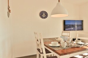 A dining area with a wooden table, white chairs, a clock, a television, and a pendant light. - Apartment Große Freiheit (Ostseebad Nienhagen)