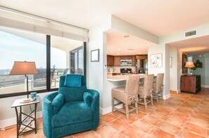 A modern living room with a teal armchair, a dining area with wicker chairs, a kitchen with a central island, and a large window offering a city view. - #306 Seychelles 2/2 Southeast Corner Oceanfront Condo on Traffic Free Beach (Daytona Beach Shores)