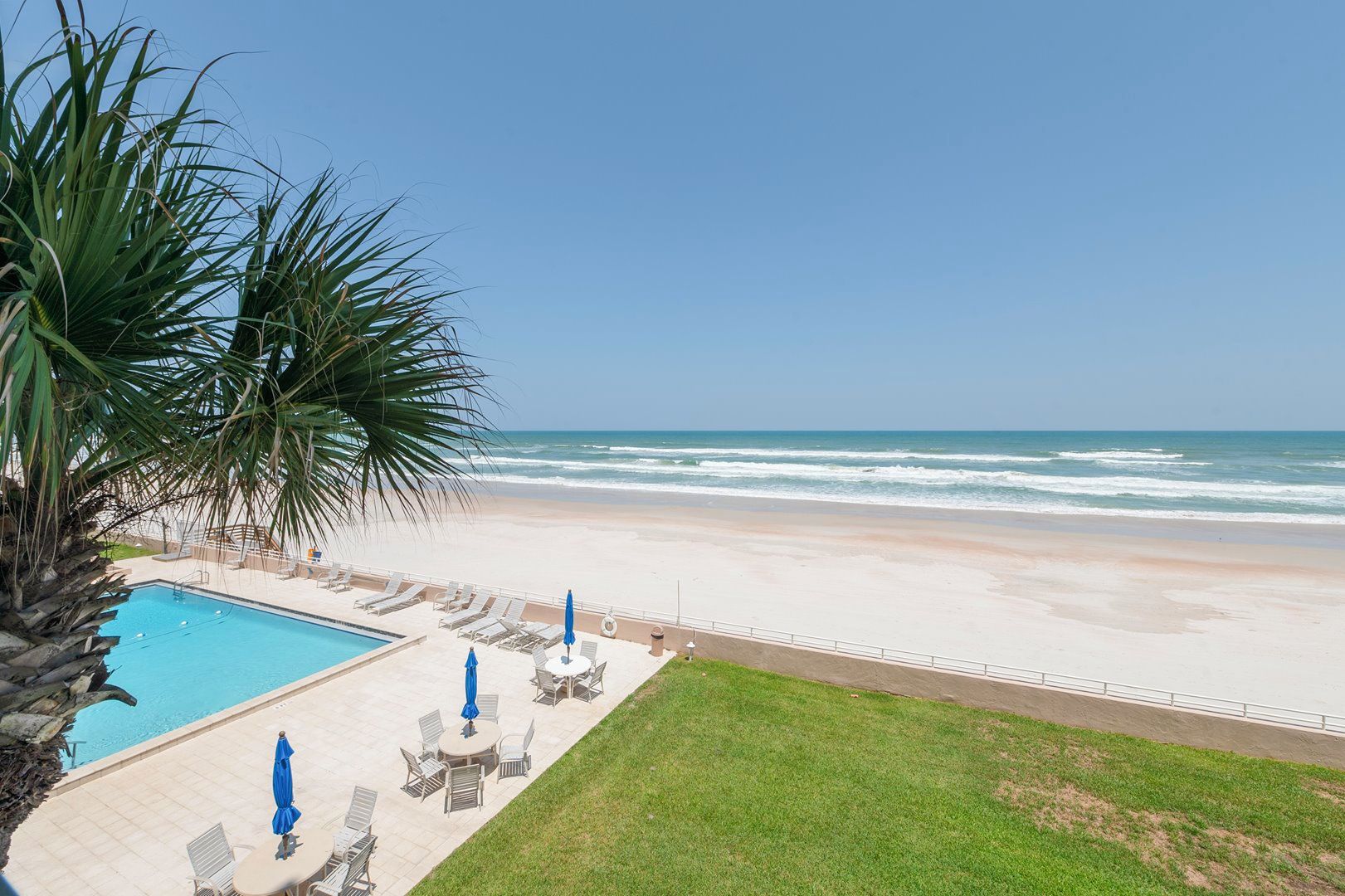 A pool area with lounge chairs and umbrellas, a beach with a clear view of the ocean, and a grassy area.