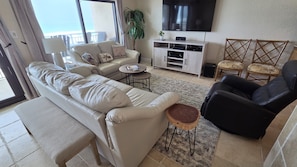 Ein Wohnzimmer mit einer Couch, einem schwarzen Sessel, einem kleinen runden Tisch, einem Fernseher und einer Pflanze.