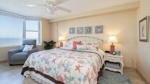 A bedroom with a bed, two bedside lamps, a chair, a window, and a wall with framed artwork. - 3202 Towers in Ponce Inlet, FL (Ponce Inlet)