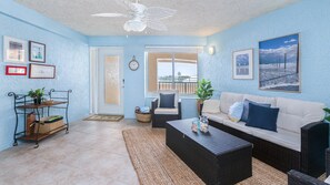 A living room with a sofa, coffee table, and a ceiling fan. There are framed pictures on the walls and a window with a view. - 3202 Towers in Ponce Inlet, FL (Ponce Inlet)