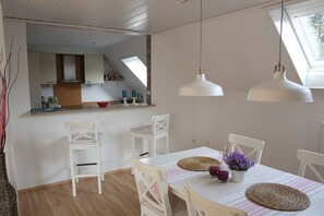 Un coin repas avec une table dressée pour deux personnes, des chaises blanches et des luminaires suspendus. On aperçoit une cuisine au fond.