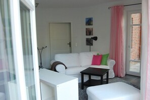 A modern living room with a white sofa, a black coffee table, and a white ottoman. There is a door leading to a balcony with pink curtains. - Buchholzer Steinbachtal (Buchholz)