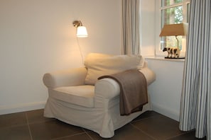 A single beige armchair with a brown throw blanket, positioned next to a window with a lamp on the wall. - Buchholzer Steinbachtal (Buchholz)
