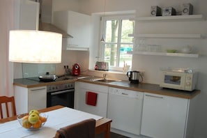 A compact kitchen with white cabinets, a stove, a sink, and a microwave. There is a window above the sink, and a bowl of fruit on the counter. - Buchholzer Steinbachtal (Buchholz)