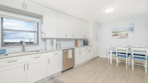 A modern kitchen with white cabinetry, a dishwasher, and a dining area with a table and chairs. - #402 Sandcastle 2/2 Oceanfront Condo Traffic Free Beach (Ponce Inlet)