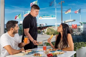 Une table mise pour le petit-déjeuner, avec trois couverts, un serveur qui sert du jus d’orange, et en arrière-plan, une vue sur la mer et des drapeaux.