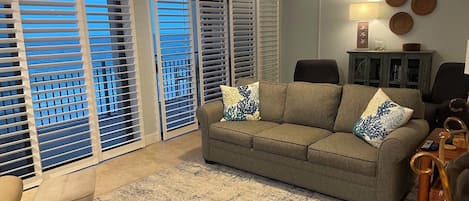 A living room with a sofa, a coffee table, a lamp, and a view through the window blinds.