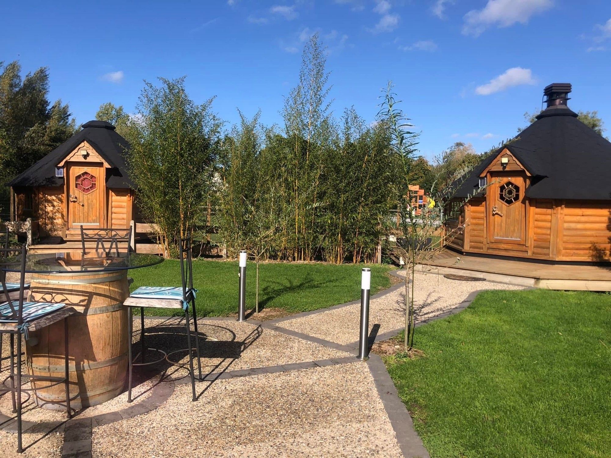 Une cabane en bois dotée d’une fenêtre hexagonale et d’une porte, entourée d’un chemin de gravier et d’une pelouse verdoyante.