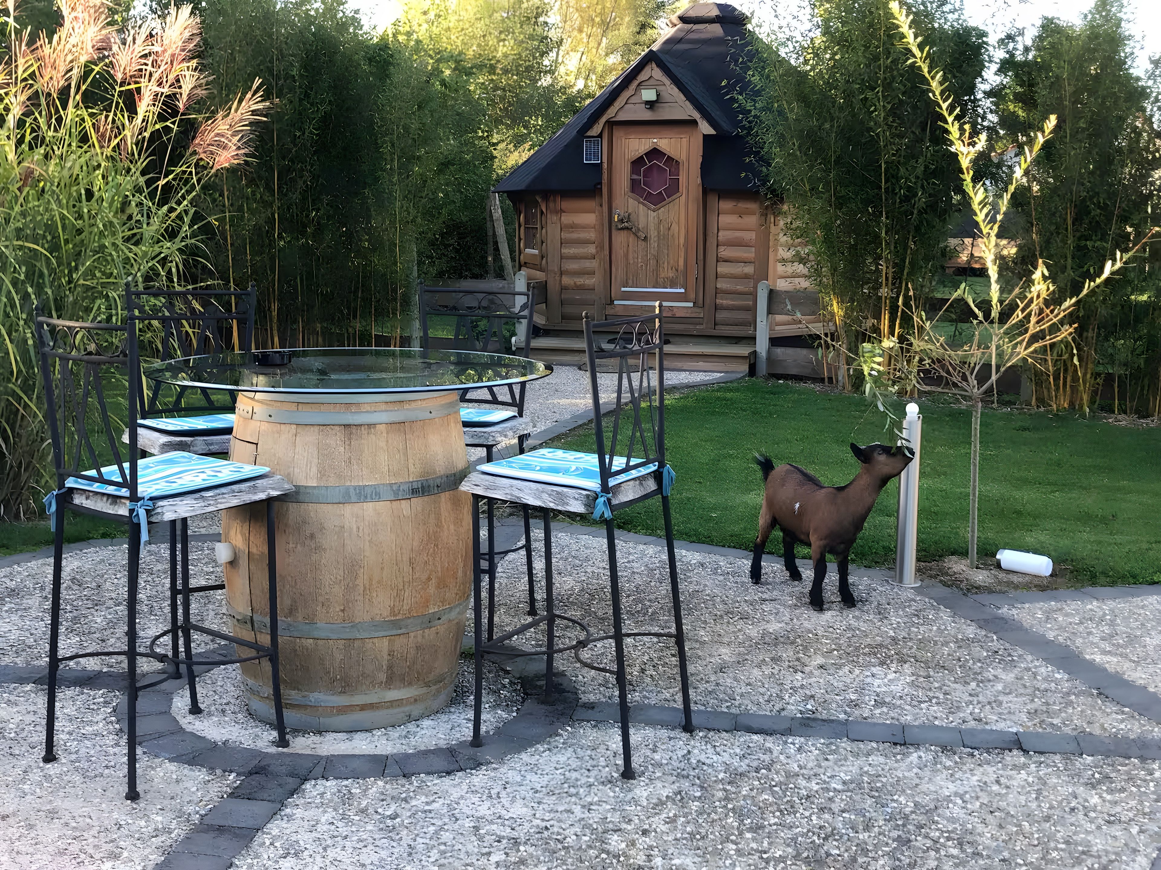Una cabaña rústica de madera con un ventanal hexagonal, rodeada por un patio de grava con sillas de metal y una mesa de barril de madera en el centro.