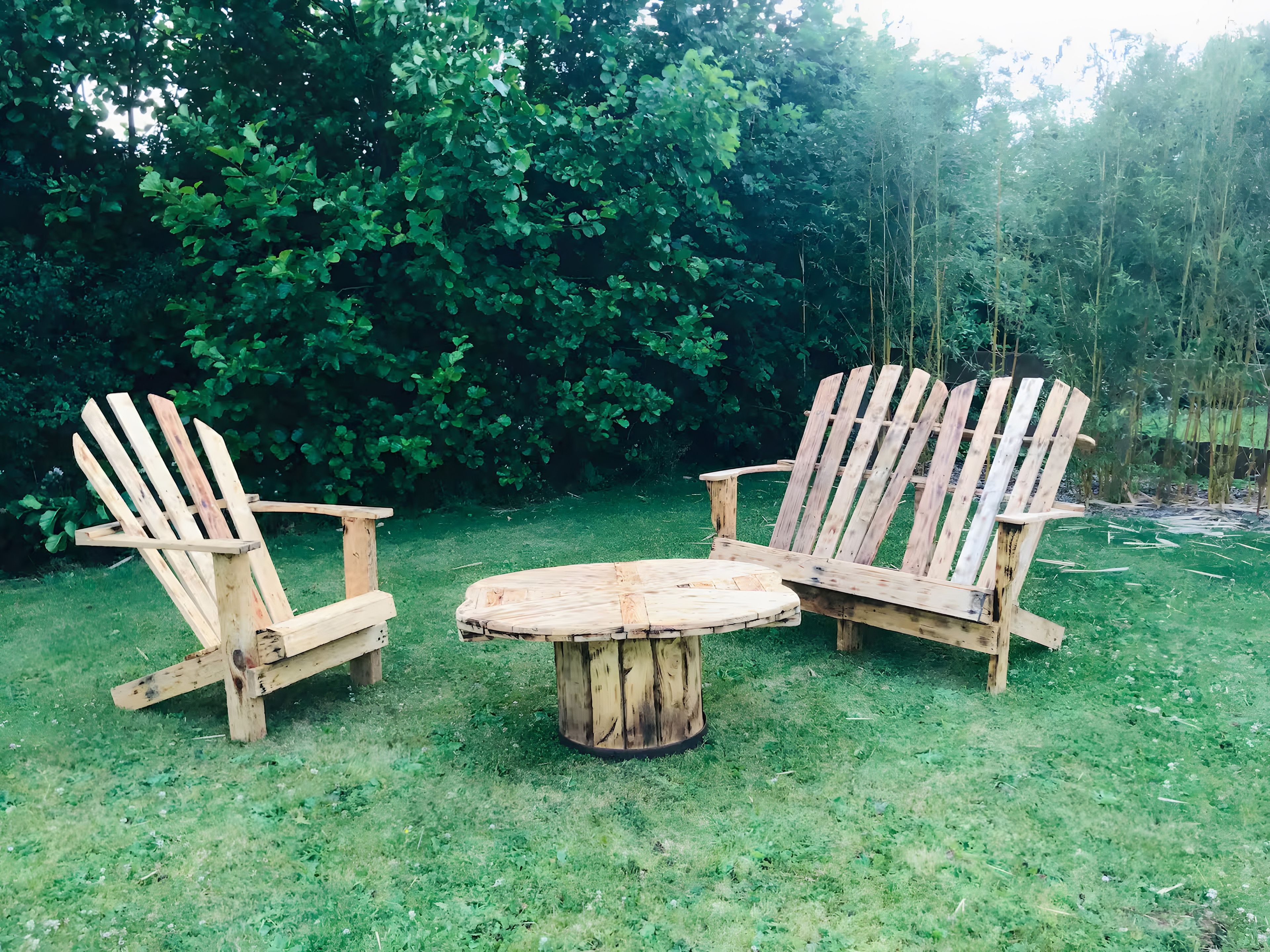 Twee Adirondack-stoelen en een houten tafel in een gazon, met weelderige beplanting op de achtergrond.