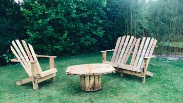 Twee Adirondack-stoelen en een houten tafel in een gazon, met weelderige beplanting op de achtergrond.