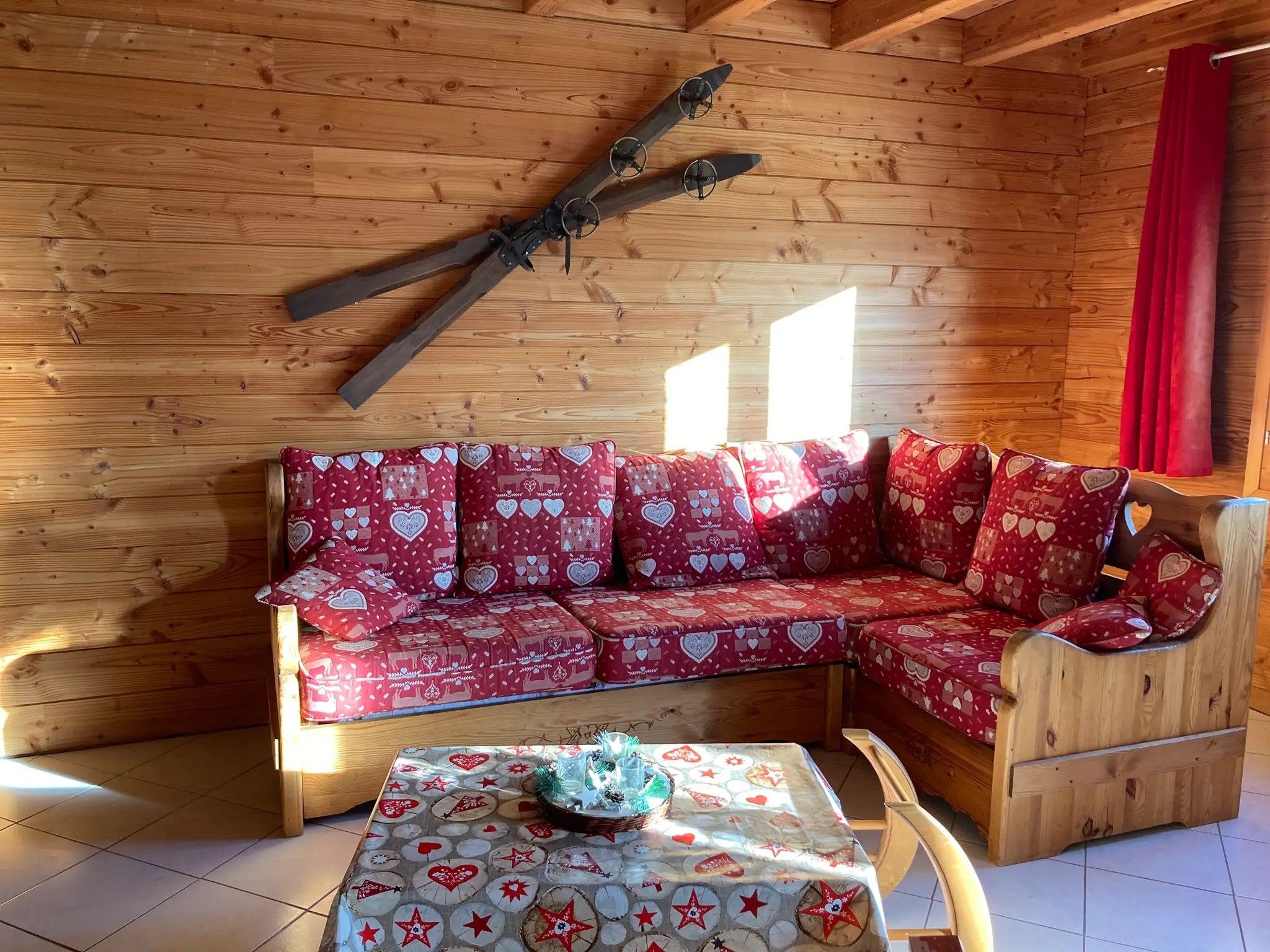 Een gezellig houten blokhutinterieur met een rode sofa met een patroon, een klein tafeltje met een feestelijk tafelkleed en een plafondventilator.