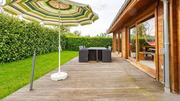 A wooden deck with a table and chairs, a striped umbrella, and a hedge in the background.