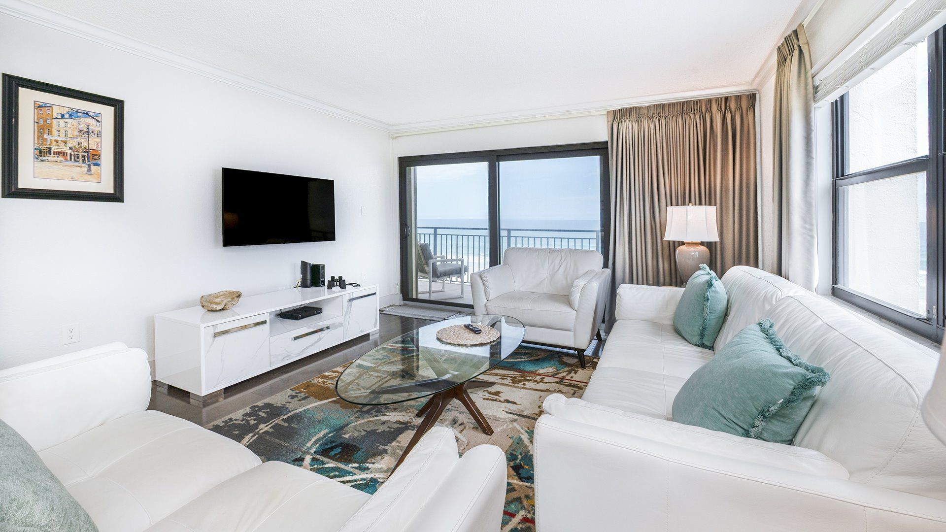 A modern hotel room with a large TV, white furniture, a glass coffee table, and a view of the sea.