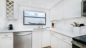 A modern kitchen with white cabinetry, a built-in dishwasher, a double sink, and a window with a view of the cityscape. - 406 Sandcastle in Ponce Inlet, FL (Ponce Inlet)
