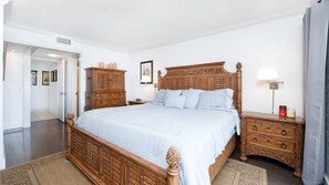 A bedroom with a wooden bed, a nightstand with a lamp, a dresser, and a door leading to another room. - 406 Sandcastle in Ponce Inlet, FL (Ponce Inlet)