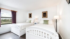 A hotel room with two single beds, a window with curtains, a nightstand with a lamp, and two framed pictures on the wall. - 406 Sandcastle in Ponce Inlet, FL (Ponce Inlet)