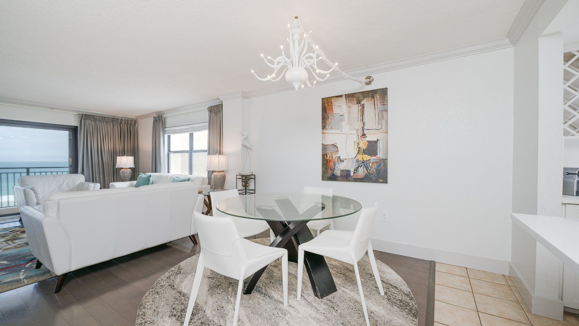 A modern living room with a glass dining table, white furniture, a painting on the wall, and a view to the outside.
