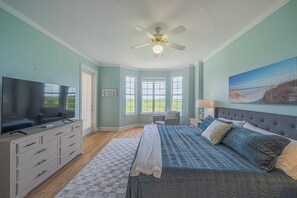 A bedroom with a large bed, a television, a ceiling fan, and a view of the outdoors through the windows. - 4404 Harbour Village (Ponce Inlet)