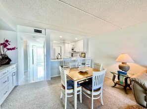 A dining area with a wooden table and white chairs, a kitchen in the background, and a lamp on a side table. - #504 Tide Water Oceanview Condo (Ponce Inlet)