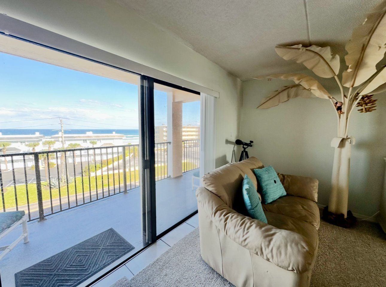 A room with a sliding glass door leading to a balcony, a beige sofa with cushions, and a large decorative plant.