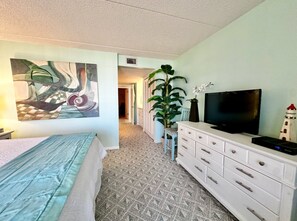Ett hotellrum med en säng, en byrå, en tv, en tavla och en växt.