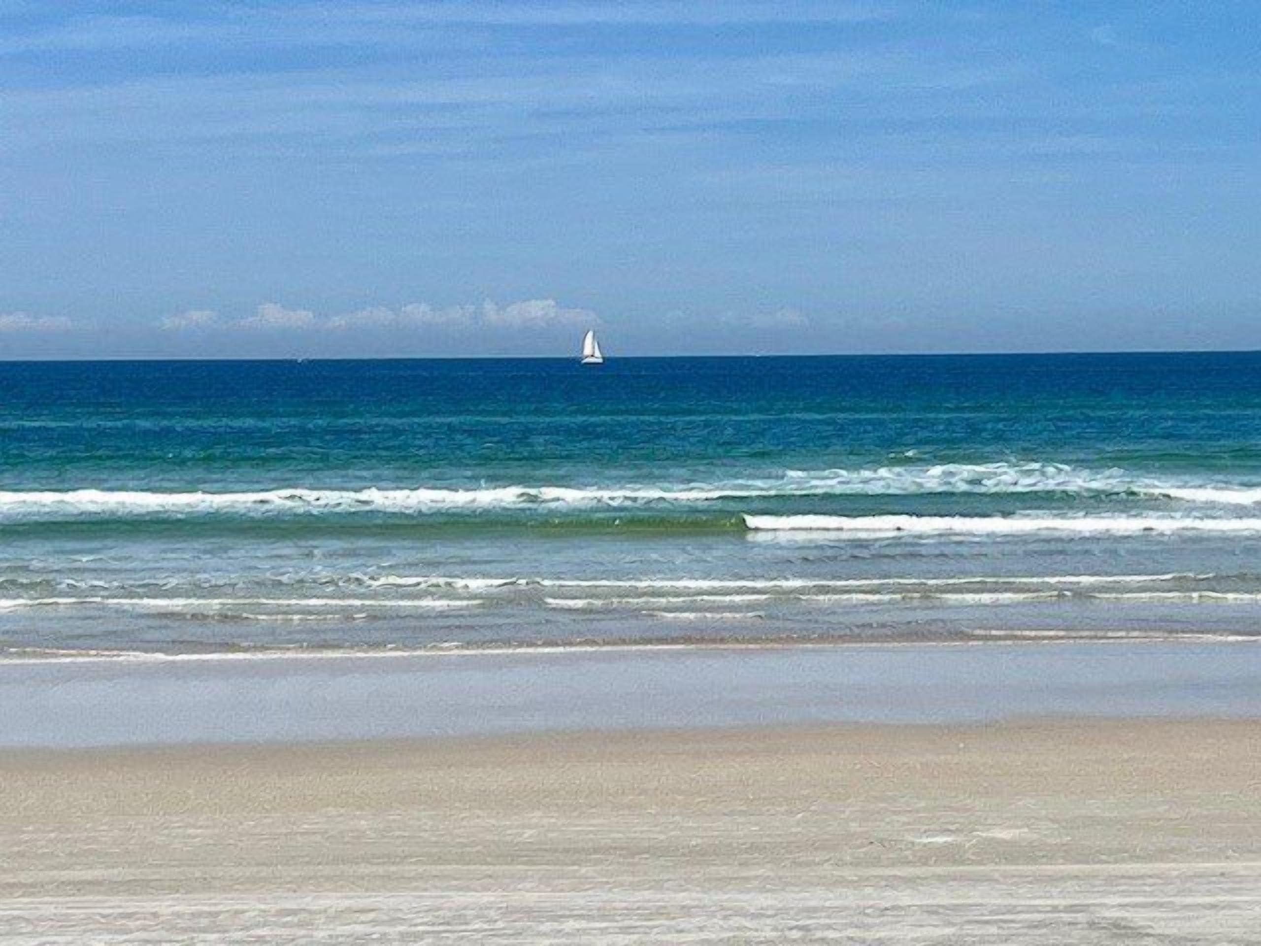 Una playa con un velero a lo lejos, mar tranquilo y cielo despejado.