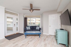 A room with a ceiling fan, a sofa with patterned cushions, a light blue dresser, and a large window with a view of buildings. - 5203 Towers in Ponce Inlet, FL (Ponce Inlet)