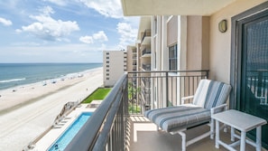 A balcony with a view of a beach and ocean, a pool, and a chair with a striped cushion. - 602 Sand Castle So in Ponce Inlet, FL (Ponce Inlet)