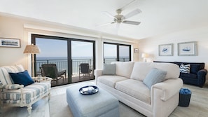 A spacious living room with a large window overlooking the ocean, a ceiling fan, and a cozy seating area with a striped armchair and ottoman. - 602 Sand Castle So in Ponce Inlet, FL (Ponce Inlet)