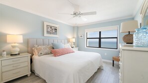 Une chambre à coucher comprenant un lit, des tables de chevet, un ventilateur de plafond, une fenêtre avec vue et un tableau encadré au mur.