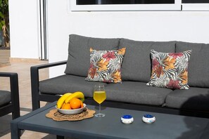 Un canapé gris agrémenté de coussins à motifs floraux, une table sur laquelle se trouve un saladier rempli de fruits et un verre contenant une boisson jaune, ainsi que deux petits objets décoratifs.