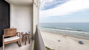A balcony with a wooden bench and a small table, overlooking a beach with a clear view of the ocean and a few people in the distance. - Seychelles 605  2/2 Direct Oceanfront condo (Daytona Beach Shores)