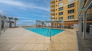 A swimming pool area with a clear view of the ocean, a covered seating area, and a multi-story building with balconies. - 606 Grand Coquina in Daytona Beach Shores, FL (Daytona Beach Shores)