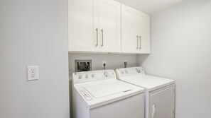 A laundry room with two white washers and dryers, a white cabinet above, and a power outlet on the wall. - 606 Grand Coquina in Daytona Beach Shores, FL (Daytona Beach Shores)