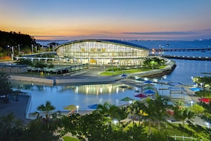 A modern building with a glass facade, a waterfront, and a pool area with umbrellas. - Central Darwin 2-Bed Apartment With Space (Darwin City)