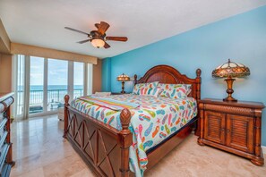Quarto com uma cama grande, cabeceira de madeira, duas luminárias de mesa e vista para o oceano através de amplas janelas.