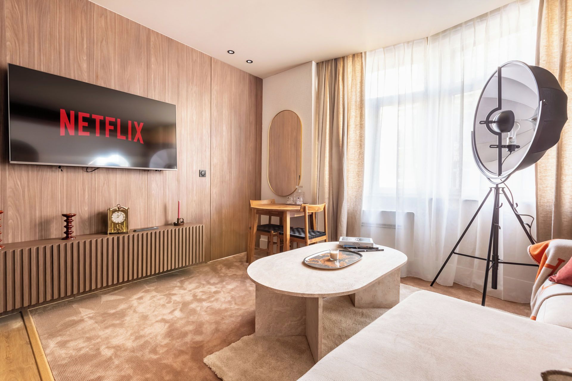 A hotel room with a television displaying 'NETFLIX,' a wooden wall, a round mirror, a small table with a tray, a tripod, and a bed.