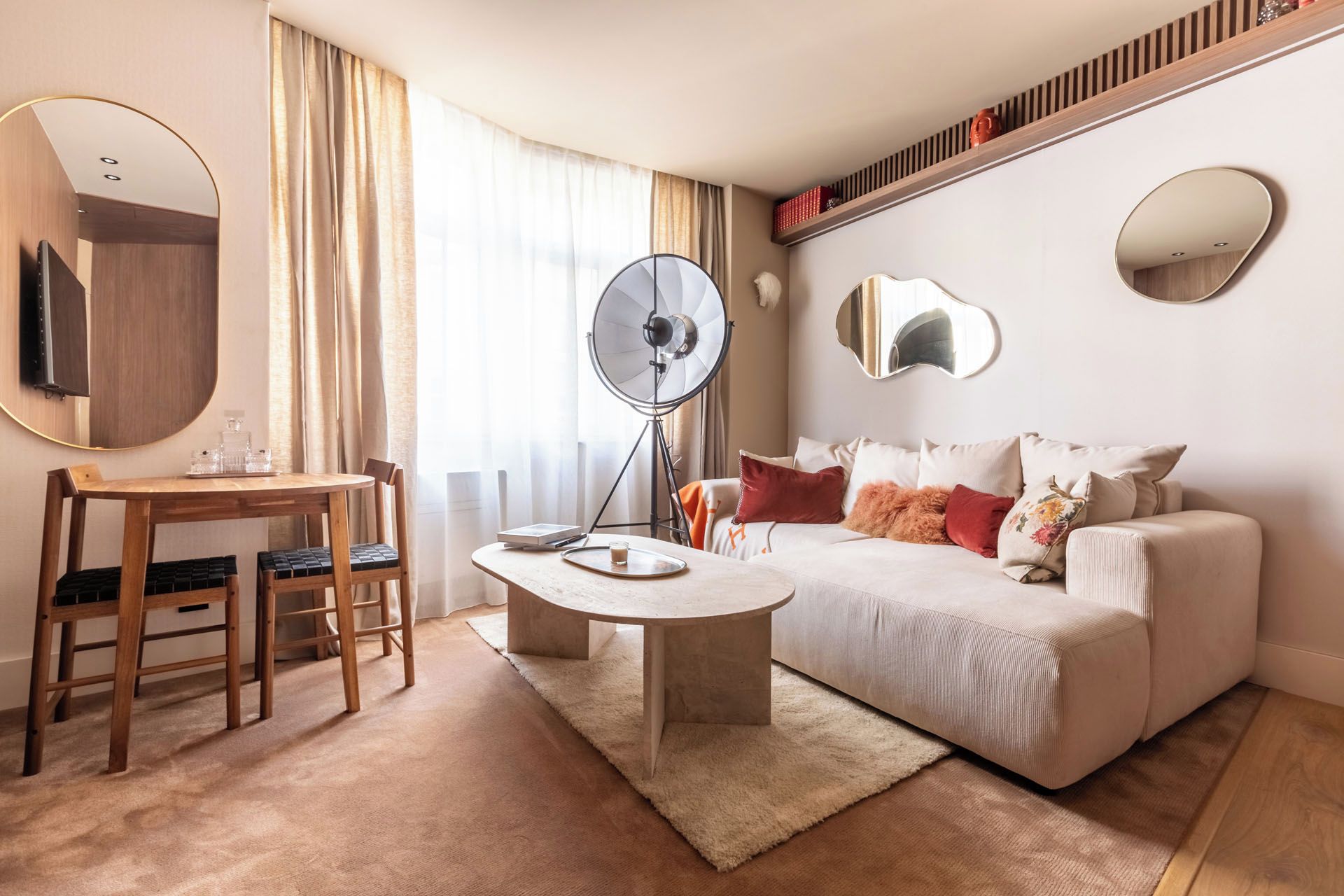 Une chambre d’hôtel moderne avec une table basse ronde, un canapé agrémenté de coussins décoratifs, un téléviseur fixé au mur et une grande fenêtre avec des rideaux.