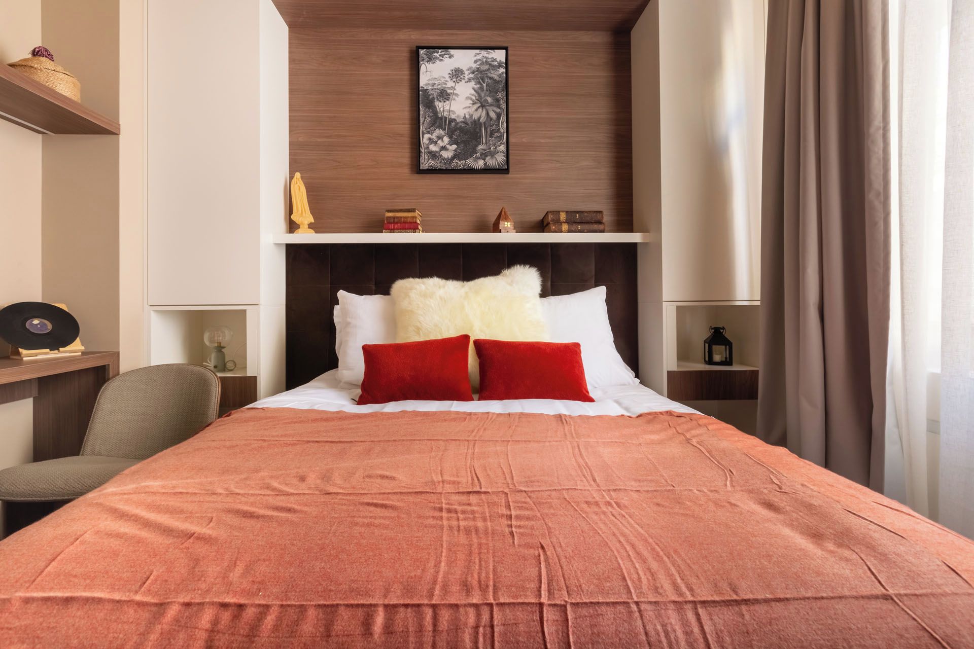 Ein Hotelzimmer mit einem Bett, einem Sessel, einem Regal mit Dekoration, einem gerahmten Bild an der Wand und einem Fenster mit Vorhängen.